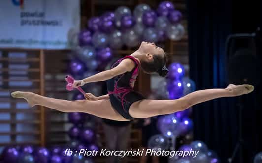 Młoda gimnastyczka w pełnym rozkroku podczas skoku trzymająca różowe pałeczki na tle balonów.