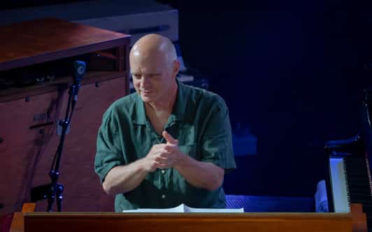 Łysy mężczyzna siedzi przy instrumencie klawiszowym na scenie i klaszcze w dłonie w niebieskim świetle.
