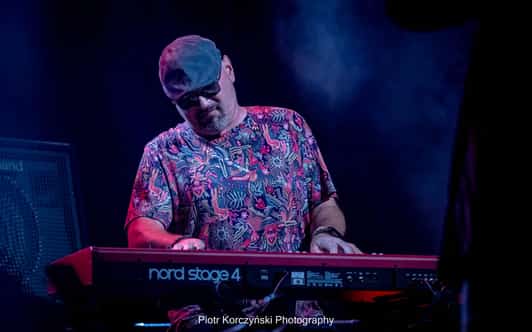 Mężczyzna w okularach przeciwsłonecznych gra na czerwonym instrumencie Nord Stage 4 na ciemnej scenie.
