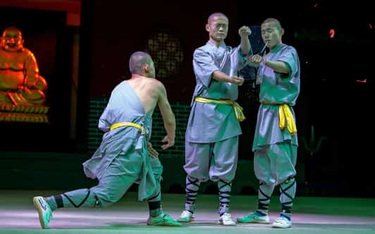 Trzech mnichów Shaolin w szarych szatach i żółtych pasach wykonuje pokaz sztuk walki na scenie przed posągiem Buddy.