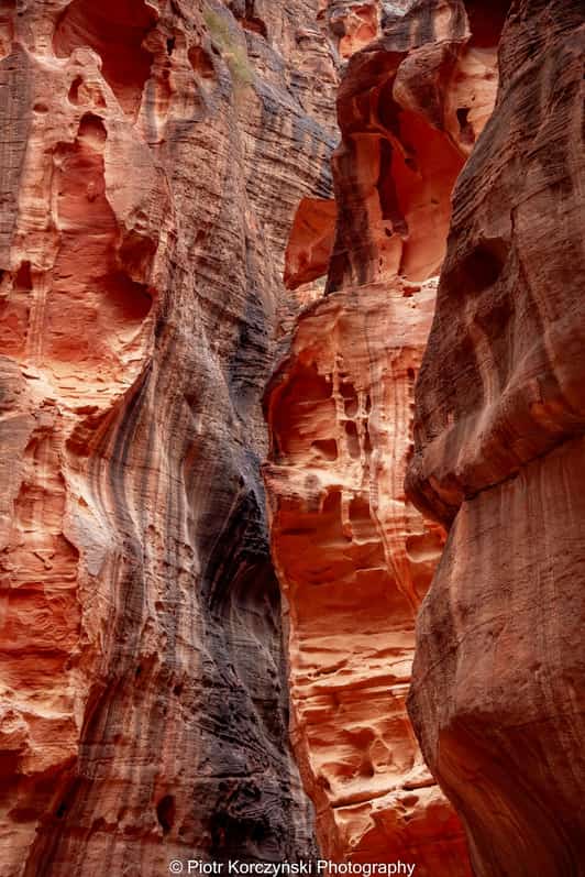 Wąski wąwóz z czerwonymi, warstwowymi ścianami skalnymi i smugami światła między szczelinami.