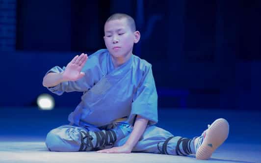 Praktykujący kung fu w szarej szacie siedzi na podłodze w rozciągniętej pozycji z uniesioną dłonią.