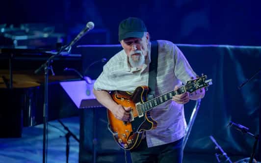 Mężczyzna w czapce gra na gitarze elektrycznej na scenie oświetlonej niebieskim światłem.