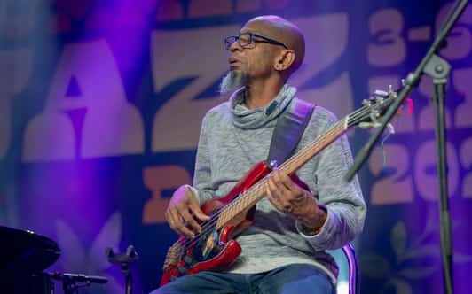 Muzyk w okularach gra na czerwonej gitarze basowej na scenie w fioletowym świetle.