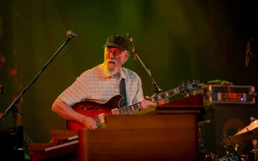 Mężczyzna w czapce gra na gitarze elektrycznej na scenie, częściowo zasłonięty klawiaturą na pierwszym planie.