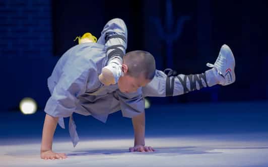 Młody mnich Shaolin utrzymuje balans na dłoniach, nogi założone za głowę podczas pokazu na scenie.