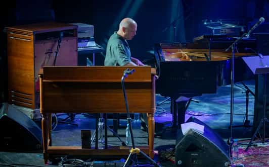 Łysy muzyk gra na fortepianie obok dużych organów na scenie skąpanej w niebieskim świetle.