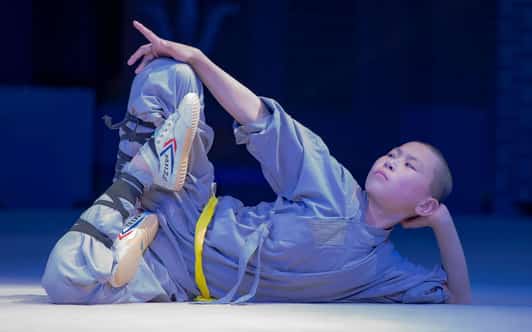 Młody mnich Shaolin w szarym habicie z żółtym pasem leżący na boku i trzymający uniesioną stopę.