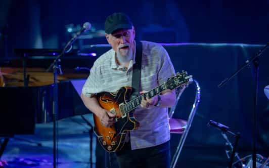 Mężczyzna w czapce gra na gitarze elektrycznej na scenie w niebieskim świetle.
