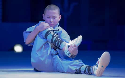 Młody mnich Shaolin w szarym stroju treningowym siedzi na scenie i trzyma zgiętą nogę podczas rozciągania.