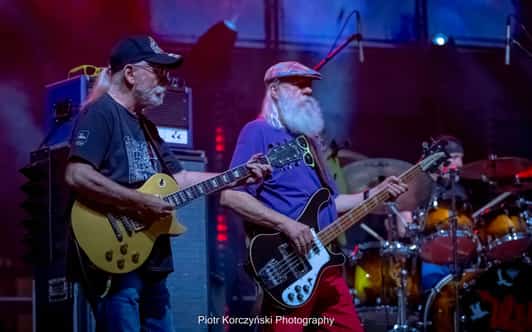 Dwóch brodatych muzyków gra na gitarze i basie na oświetlonej scenie, a perkusista siedzi w tle.