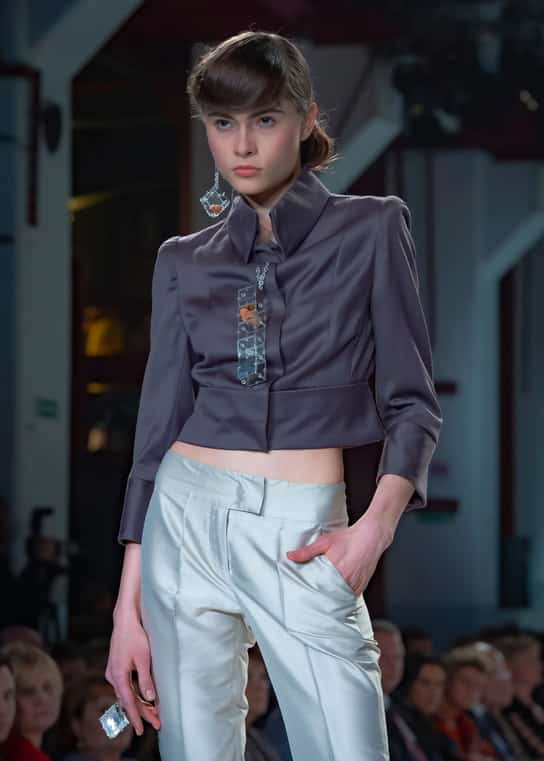 Modelka w krótkiej ciemnej marynarce i jasnych spodniach maszeruje po wybiegu, dłoń w kieszeni, widownia w tle.