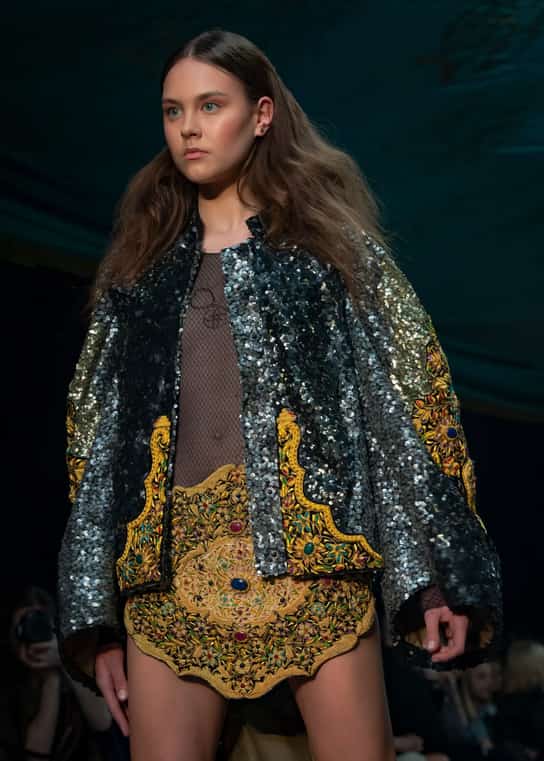 Modelka na wybiegu w oversize'owej cekinowej kurtce, prześwitującej bluzce i bogato zdobionej złotej mini spódnicy.