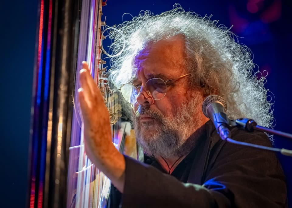 Older man with curly gray hair and glasses playing a harp by a microphone on stage.