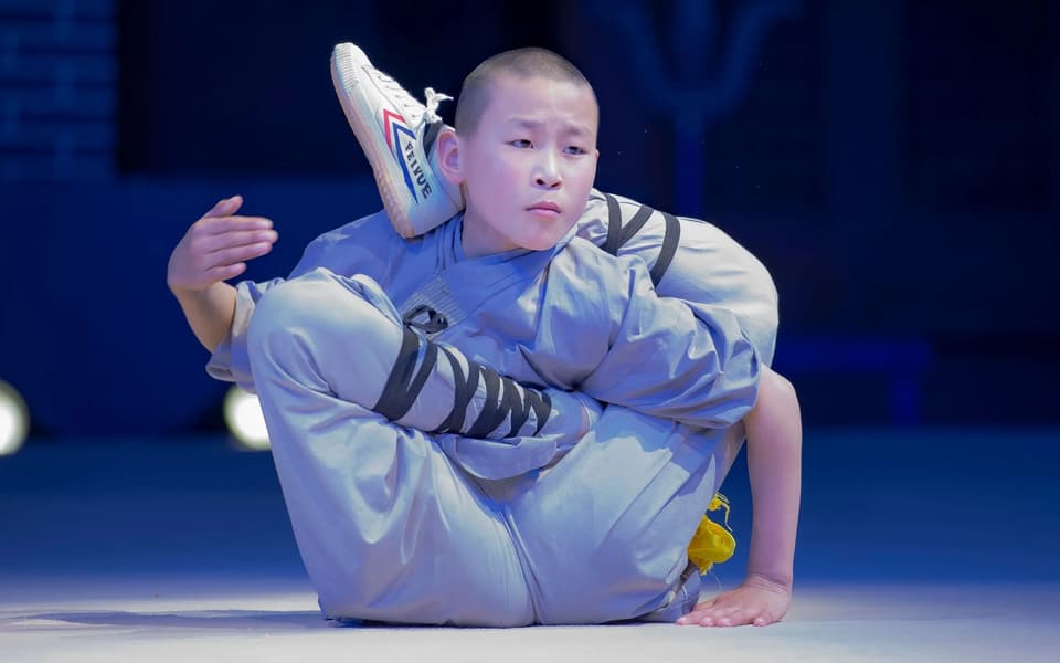Young Shaolin monk in a gray robe performing an acrobatic contortion pose on stage.