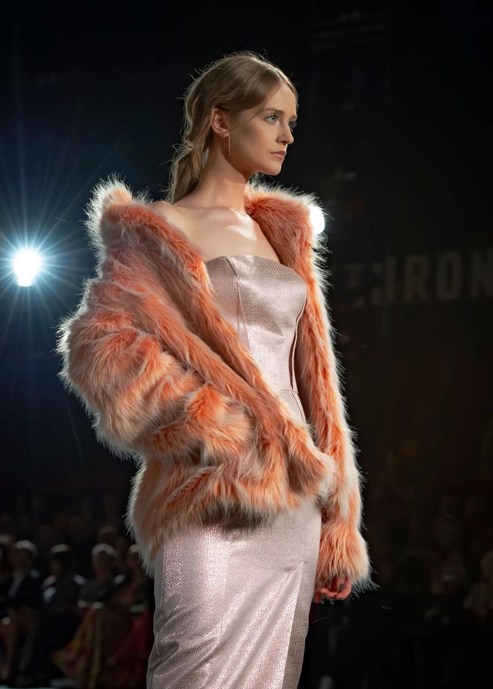 Model wearing a shimmering strapless gown and orange fur coat walks the runway, with spotlights and audience in the background.