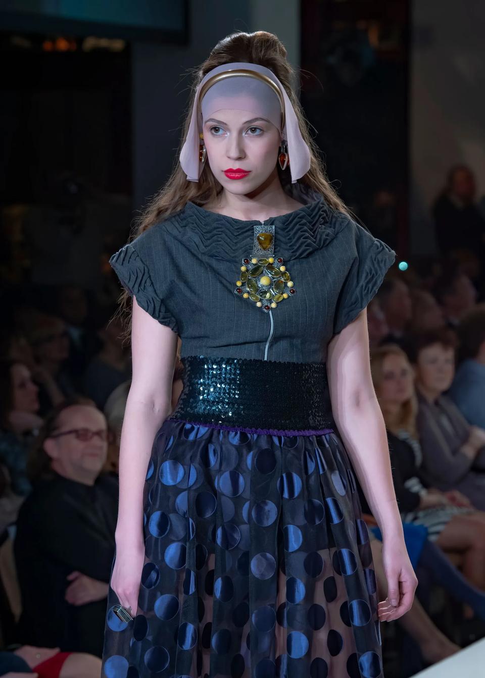 Model walks the runway in a headpiece, dark top with a large brooch and a shimmering polka-dot skirt.