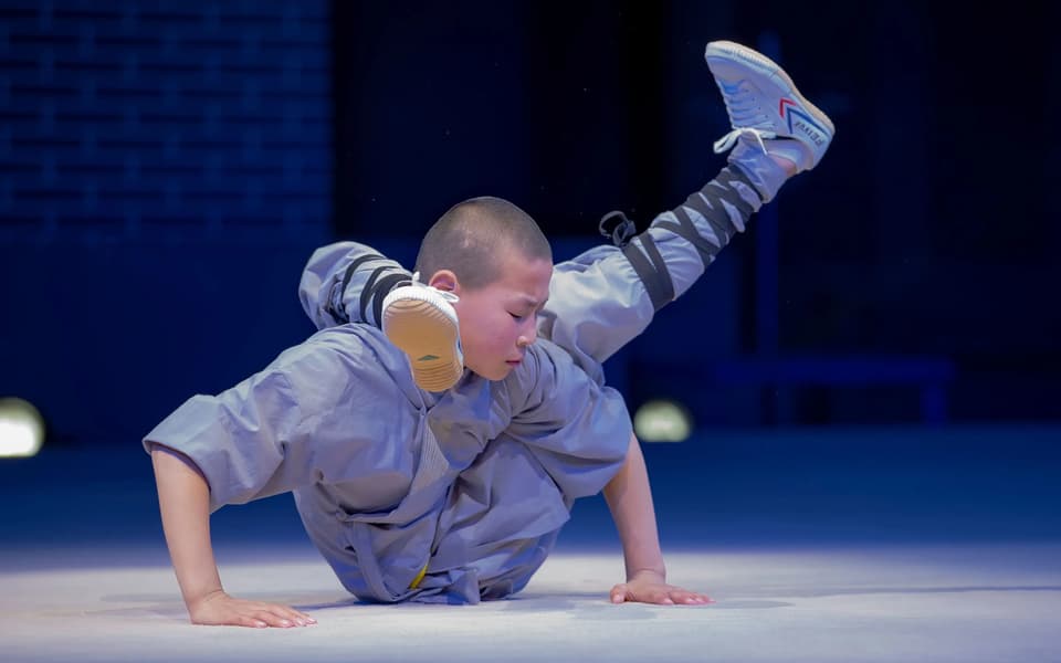 Young monk in a grey robe balances in an acrobatic handstand-like pose on a stage.
